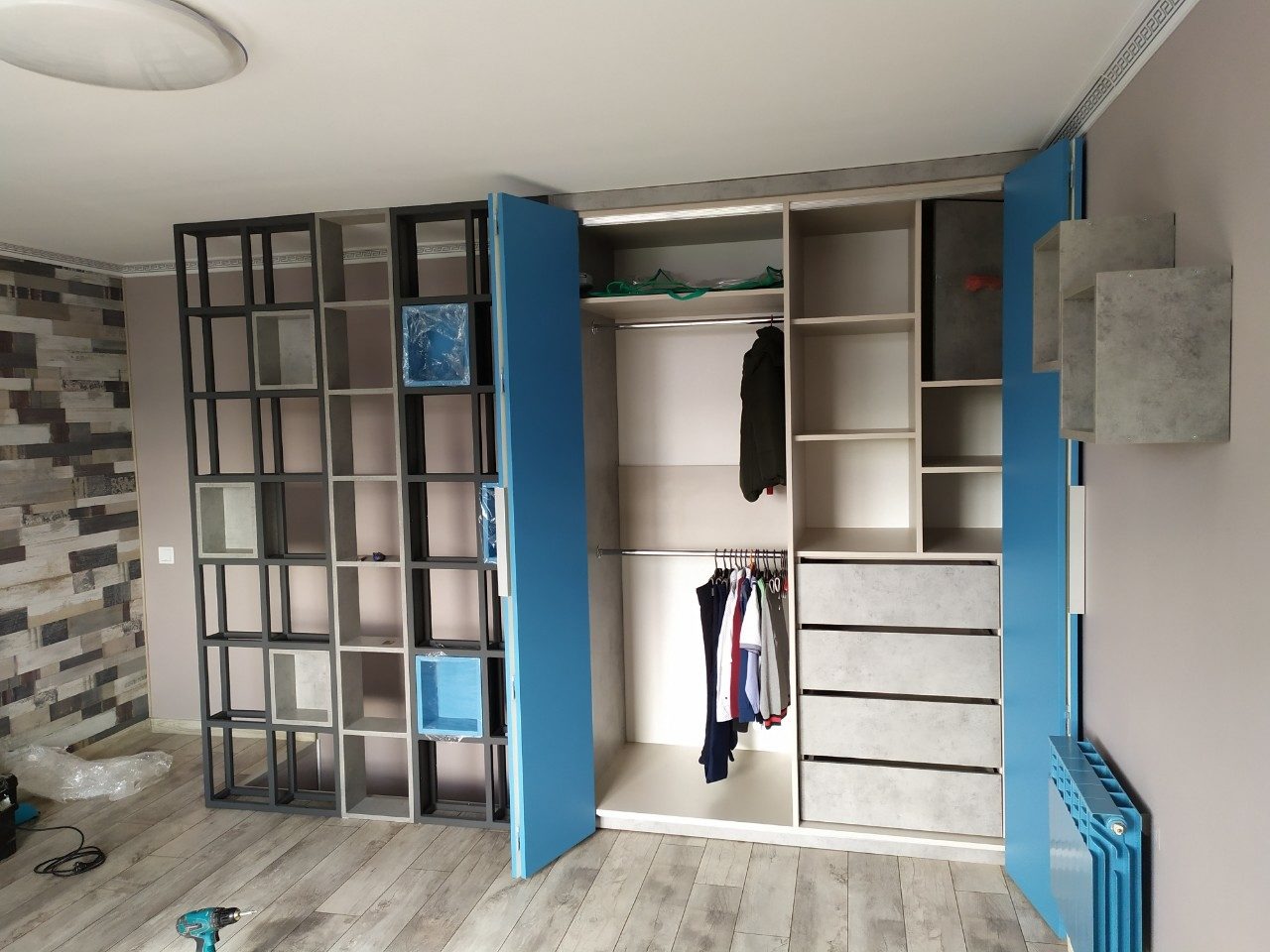 Nowoczesna szafa wnękowa z otwartymi niebieskimi drzwiami i regałem z kwadratowymi półkami, w tle ściana z dekoracyjną okładziną, na podłodze wiertarka akumulatorowa.