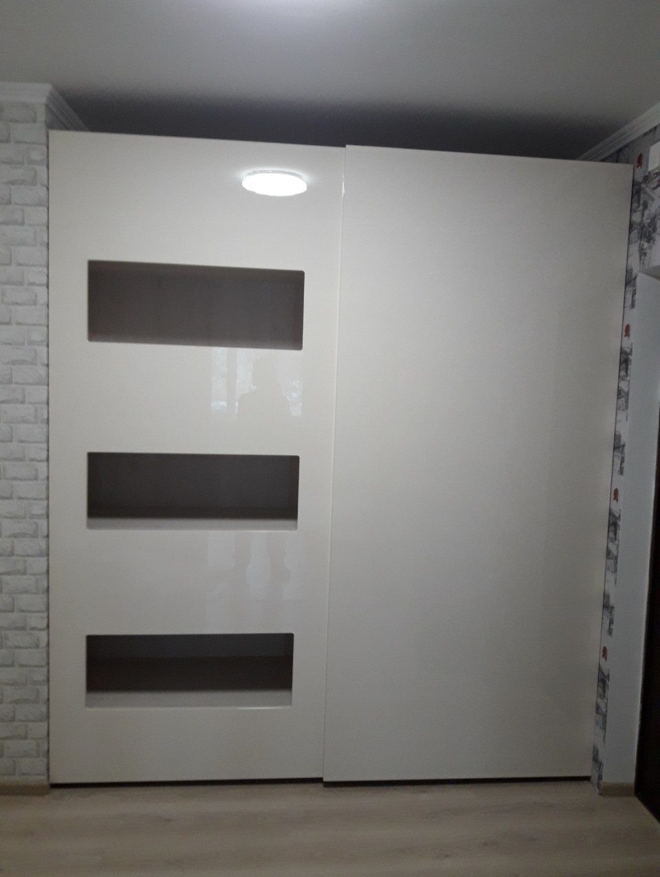 Biała szafa przesuwna do przedpokoju z trzema poziomymi, prostokątnymi wstawkami z ciemnego szkła, oświetlona od góry, ustawiona na tle ściany wykończonej białą cegłą.