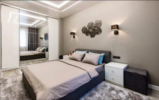 Sypialnia z łóżkiem, szafkami nocnymi i dużą szafą przesuwną z lustrzanymi drzwiami, odbijającymi fragment pokoju, oświetlona lampami ściennymi i sufitowym oświetleniem LED.