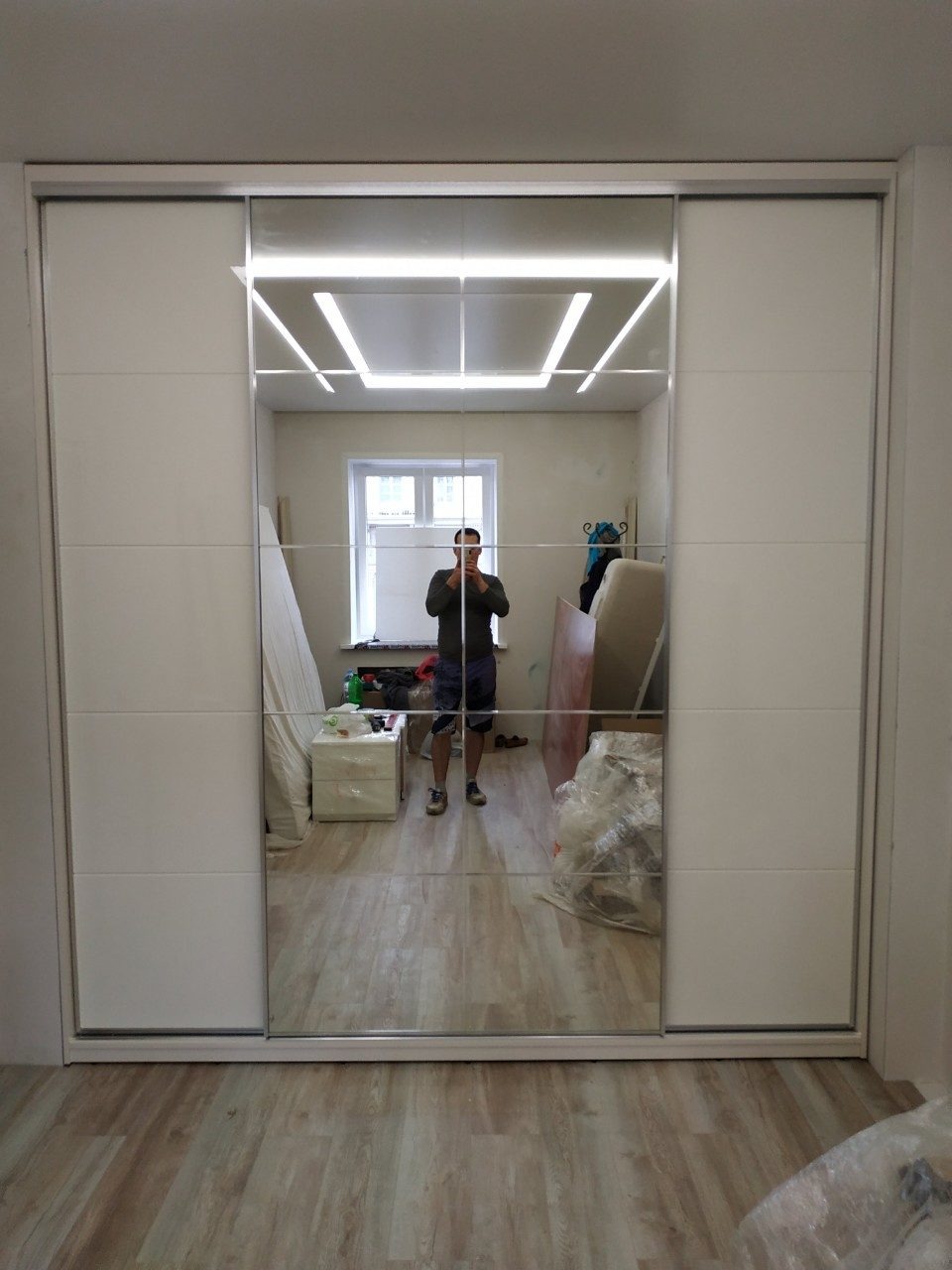 Biała szafa przesuwna z lustrzanymi drzwiami, odbijająca wnętrze pokoju z oknem i elementami wyposażenia w trakcie montażu, widoczne narzędzia i materiały budowlane.