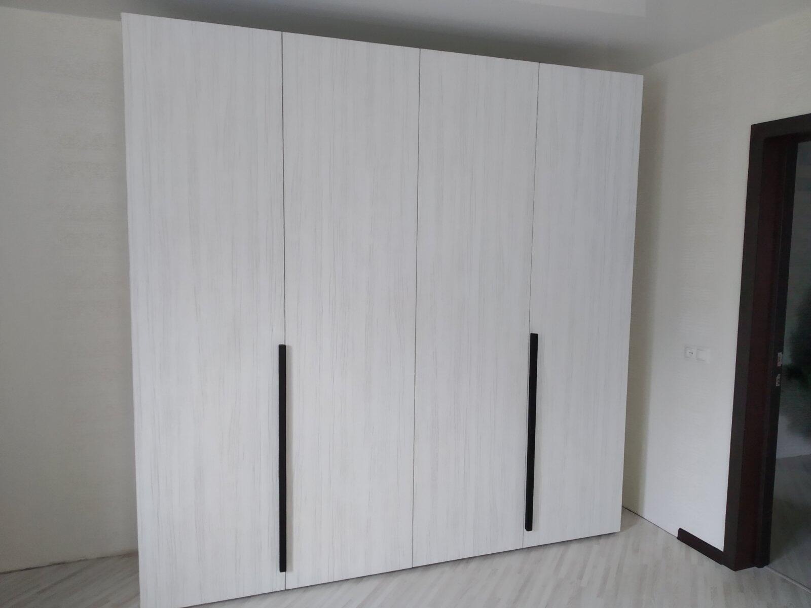 Czterodrzwiowa szafa wnękowa z białego drewna z wyraźnym rysunkiem słojów i długimi, pionowymi, czarnymi uchwytami, dopasowana do jasnej podłogi i ściany, widoczna rama drzwi po prawej stronie.