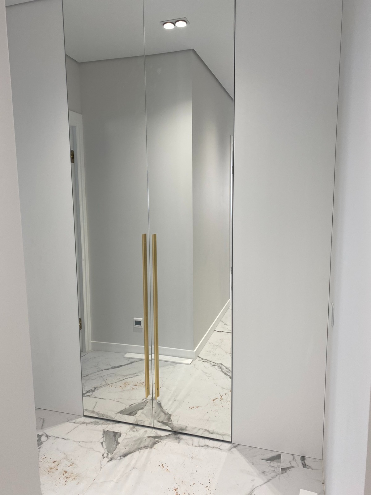 Biała szafa wnękowa z lustrzanymi drzwiami i złotymi uchwytami, odbijająca marmurową podłogę i fragment wnętrza z oświetleniem punktowym.