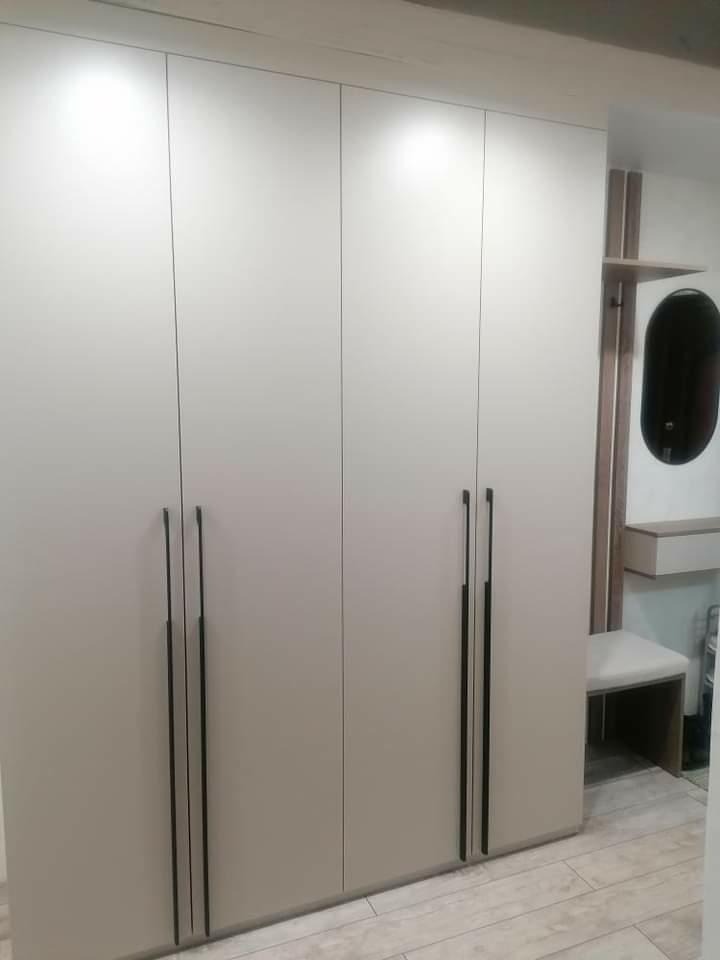 Czterodrzwiowa szafa w zabudowie w odcieniach szarości z długimi, pionowymi uchwytami, obok wnęka z owalnym lustrem i siedziskiem.