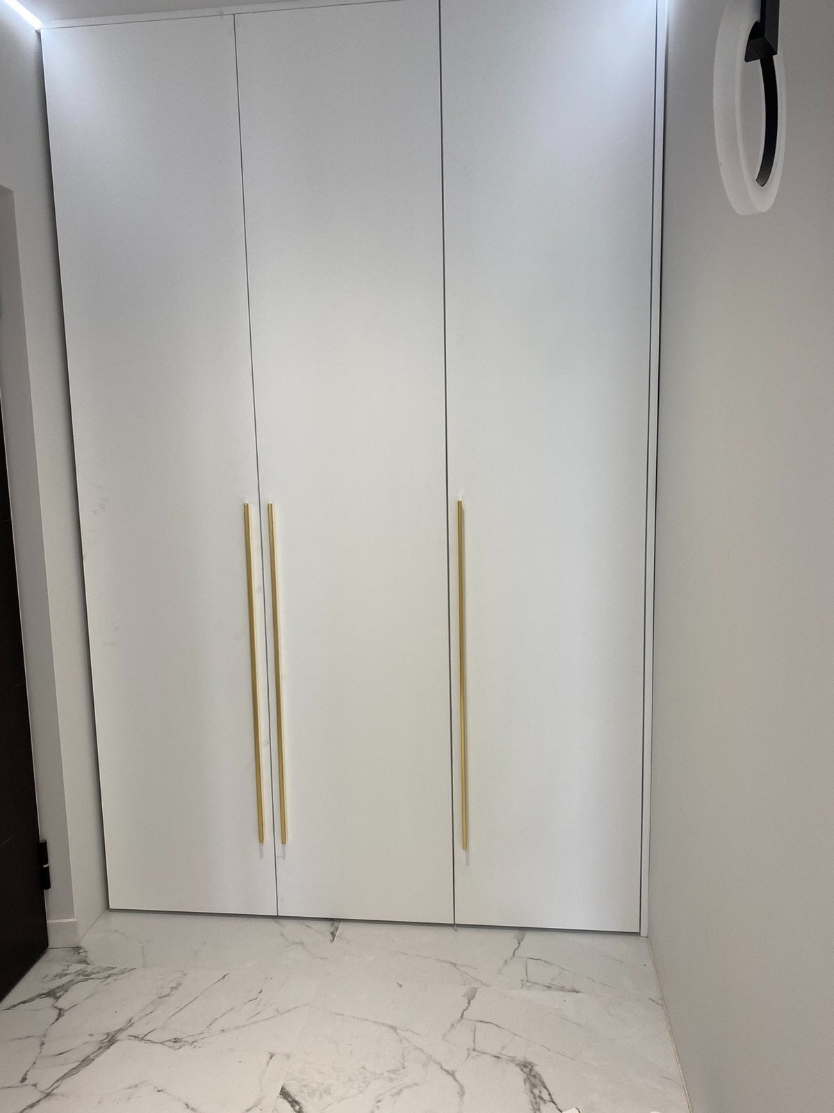 Biała, trzydrzwiowa szafa wnękowa z długimi, złotymi uchwytami na tle marmurowej podłogi i minimalistycznego oświetlenia.