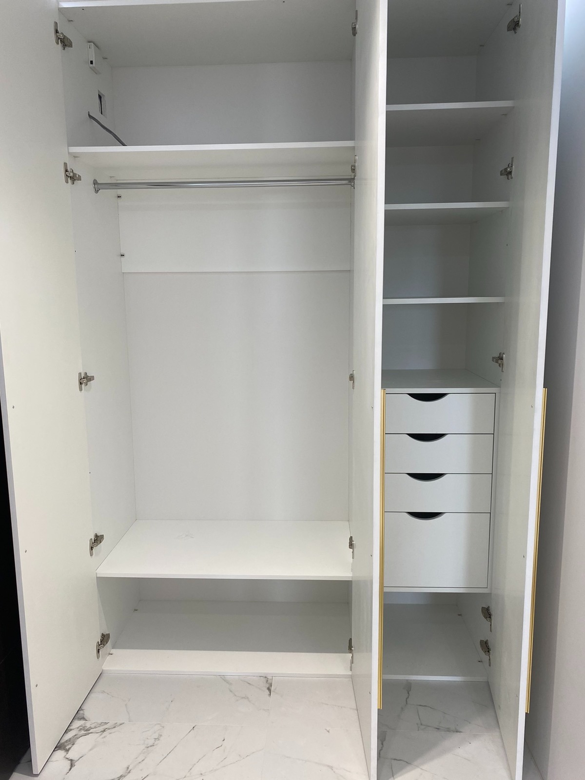 Wnętrze białej, otwartej szafy z półkami, drążkiem na ubrania i czterema szufladami, z widocznymi zawiasami i marmurową podłogą w tle.