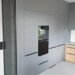 Nowoczesna, szara zabudowa kuchenna z piekarnikiem, mikrofalówką i przeszkloną witryną, z drewnianymi elementami wyspy kuchennej.