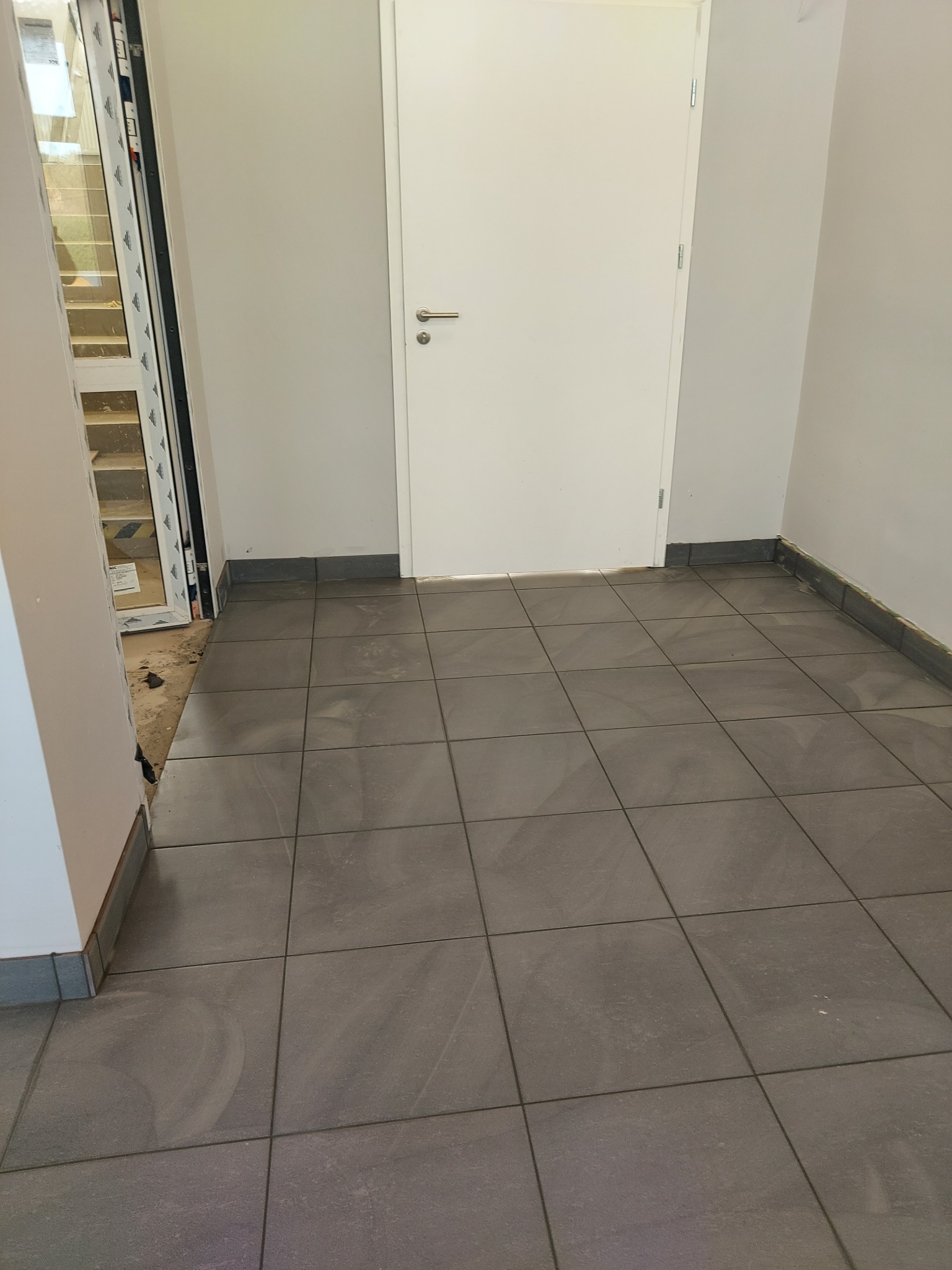 Ułożona, szara, kwadratowa podłoga z płytek ceramicznych w korytarzu z białą ścianą i drzwiami, widoczny fragment okna.