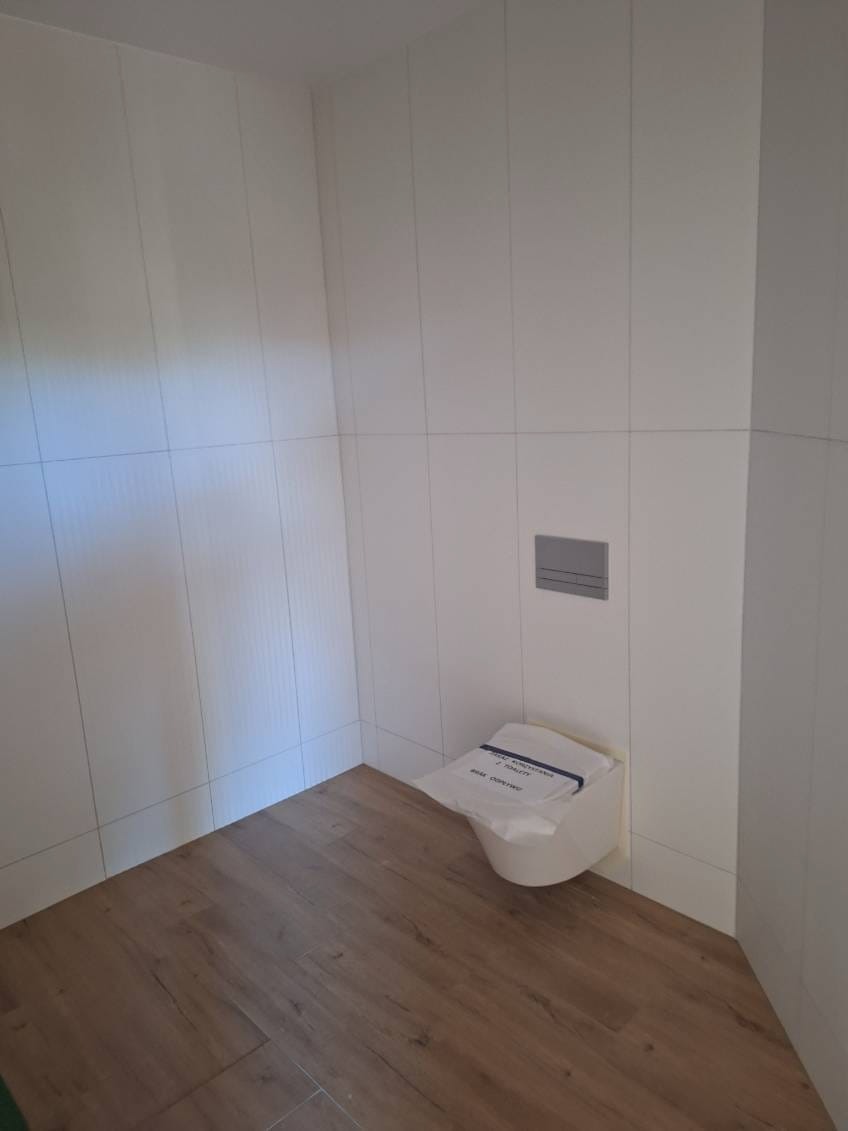 Nowoczesna, minimalistyczna toaleta z drewnopodobną podłogą i białymi, kwadratowymi płytkami na ścianach, zabezpieczona folią ochronną przed pierwszym użyciem.