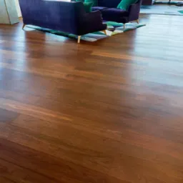 Świeżo ułożony parkiet z ciemnego drewna z widocznymi słojami, odbijający światło, z fragmentem fioletowej sofy na złotych nóżkach i wzorzystym dywanem w tle.