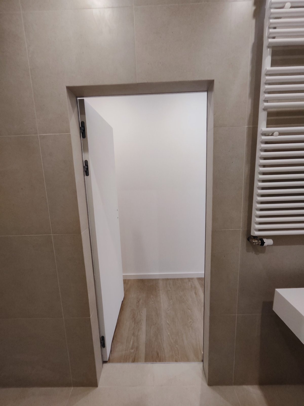 Wykończona łazienka z beżowymi płytkami, białą, częściowo otwartą ścianką działową i grzejnikiem drabinkowym po prawej stronie.