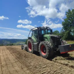 Zielony traktor Fendt z czerwonymi felgami na polu po orce, w tle pagórkowaty krajobraz pod błękitnym niebem z obłokami.