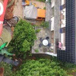 Wycinka,Pielegnacja Drzew Piotr Bogdanik - Widok z góry na podwórko z drzewami, meblami ogrodowymi i budynkiem, z perspektywy osoby wspinającej się na drzewo, widoczne liny i sprzęt wspinaczkowy, w tle maszyna do rozdrabniania gałęzi...