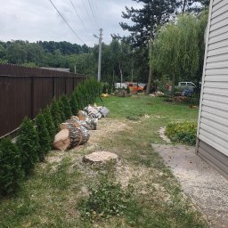 Wycinka,Pielegnacja Drzew Piotr Bogdanik - Świeżo ścięte klocki drewna ułożone rzędem wzdłuż żywopłotu z tui, pozostałości po pniu i rozsypane trociny na trawniku w ogrodzie po zakończonych pracach.