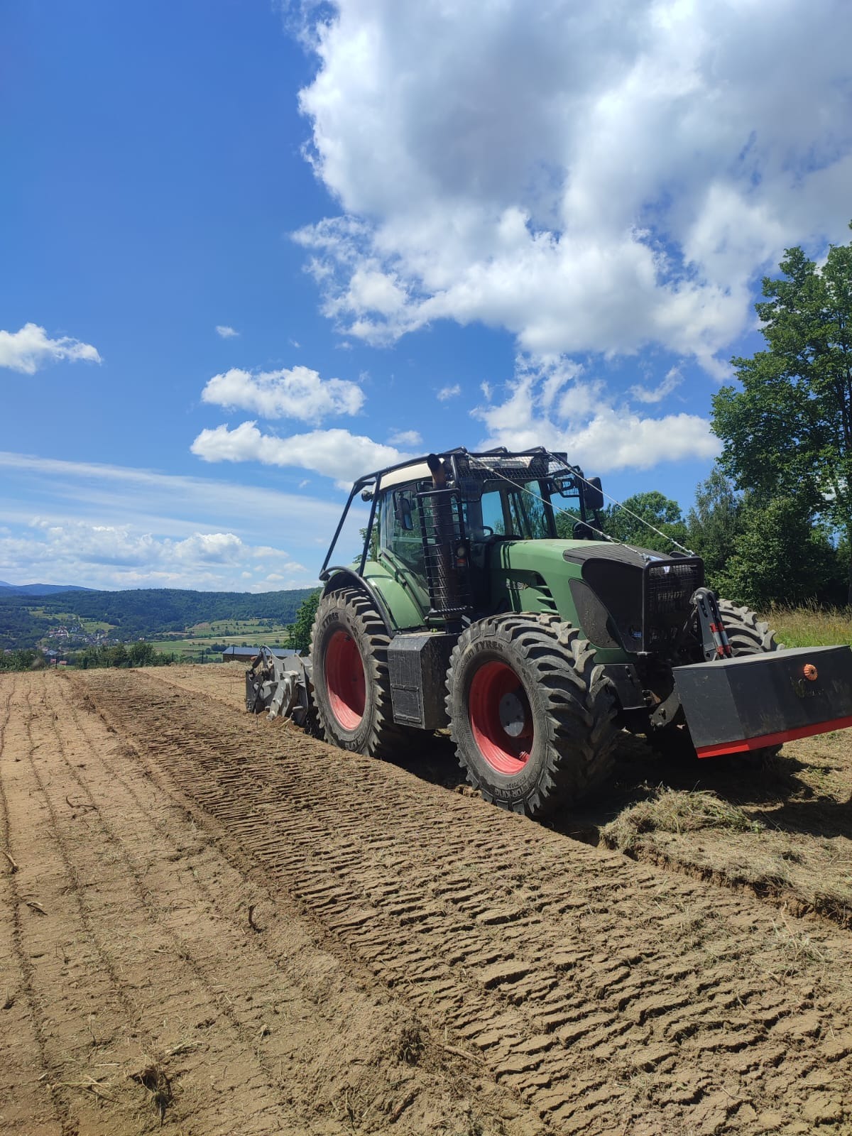 Zielony traktor Fendt z czerwonymi felgami na polu po orce, w tle pagórkowaty krajobraz pod błękitnym niebem z obłokami.