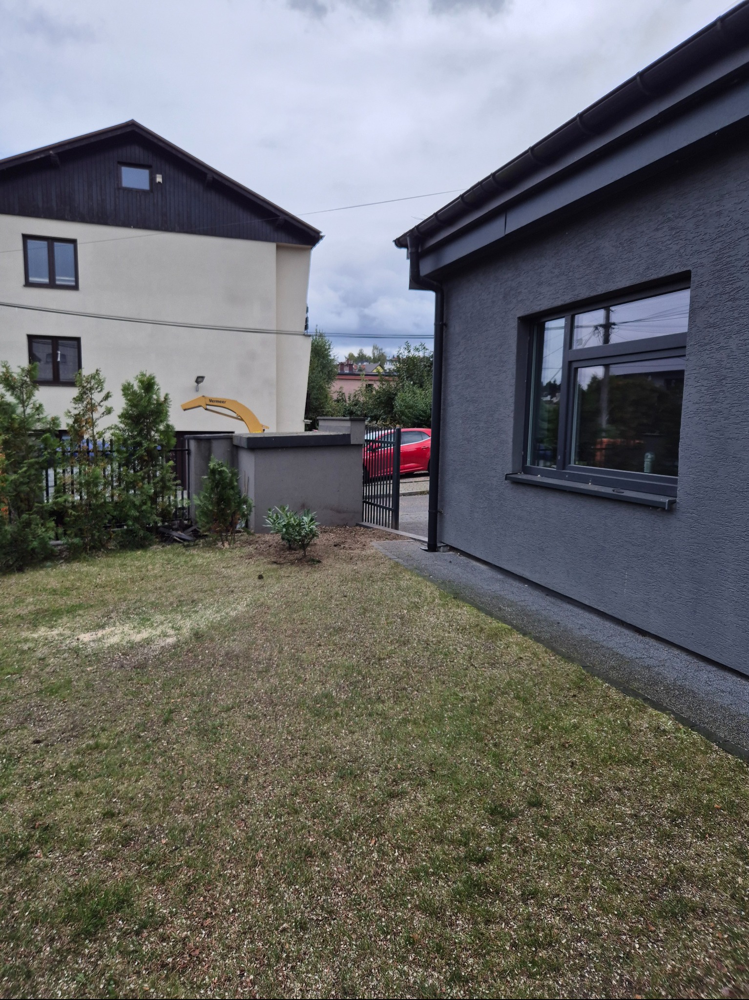 Ogród z widokiem na domy, gdzie na elewacji jednego z nich zamontowane jest ramię podnośnika koszowego. Widoczne tuje i trawnik.