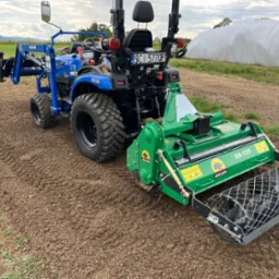 Niebieski traktor z zielonym glebogryzarką marki Mikann na świeżo przygotowanej ziemi, widok z tyłu, w tle zielone pola i biały tunel foliowy.