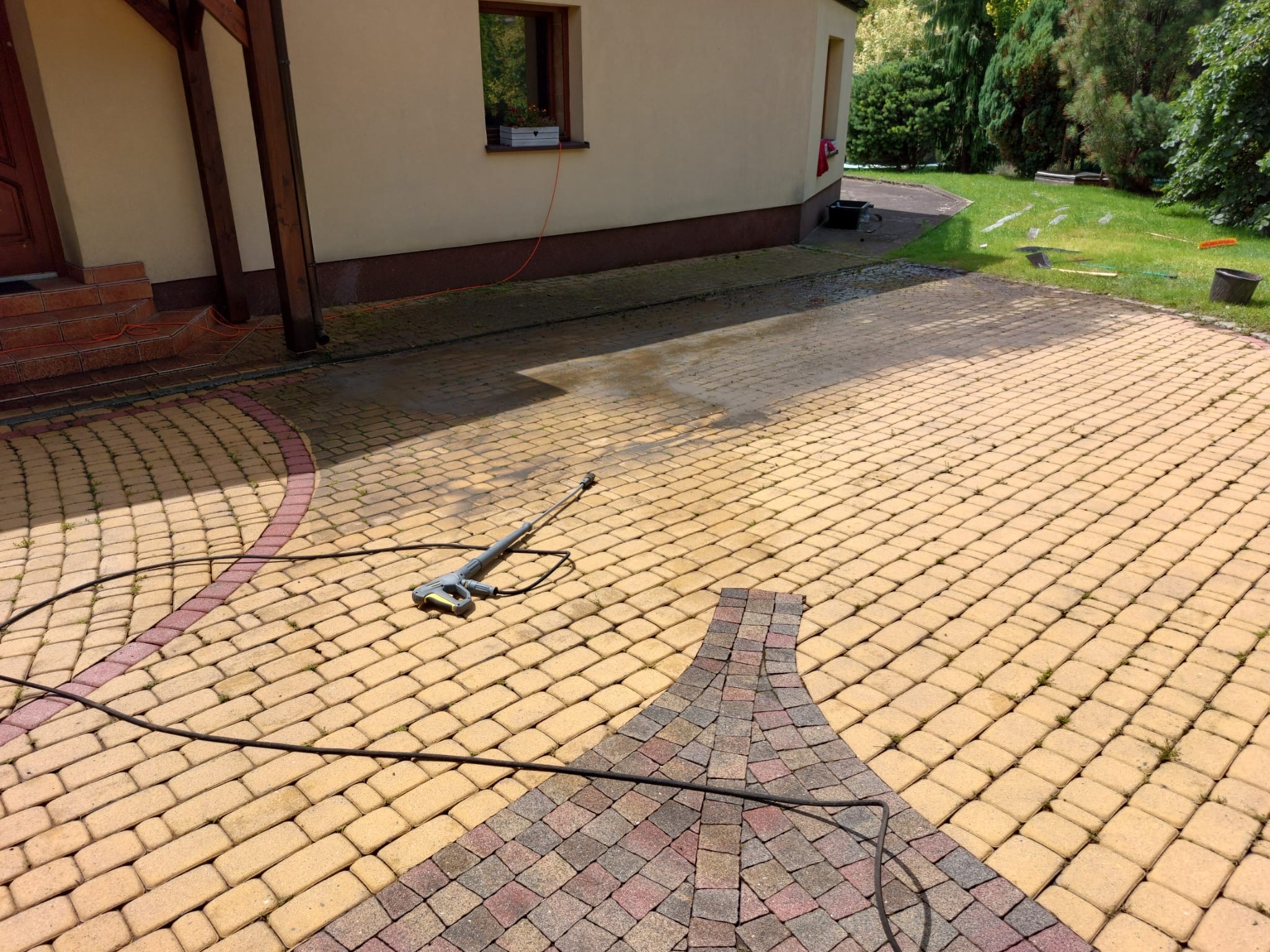 Mycie kostki brukowej na podjeździe domu za pomocą myjki ciśnieniowej. Widoczny żółty bruk, fragment domu z oknem i zielony trawnik w tle.