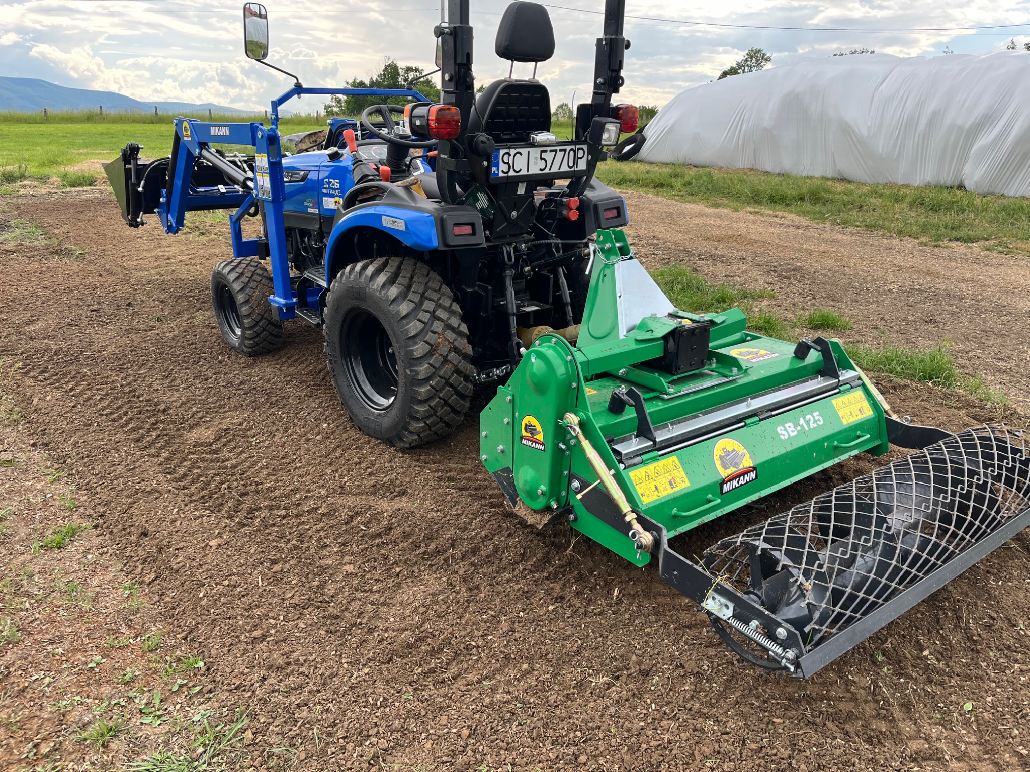 Niebieski traktor z zielonym glebogryzarką marki Mikann na świeżo przygotowanej ziemi, widok z tyłu, w tle zielone pola i biały tunel foliowy.