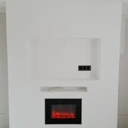 Biała ściana z wnękami i elektrycznym kominkiem z czarną obudową, widoczne gniazdka elektryczne i fragment kabla w półce