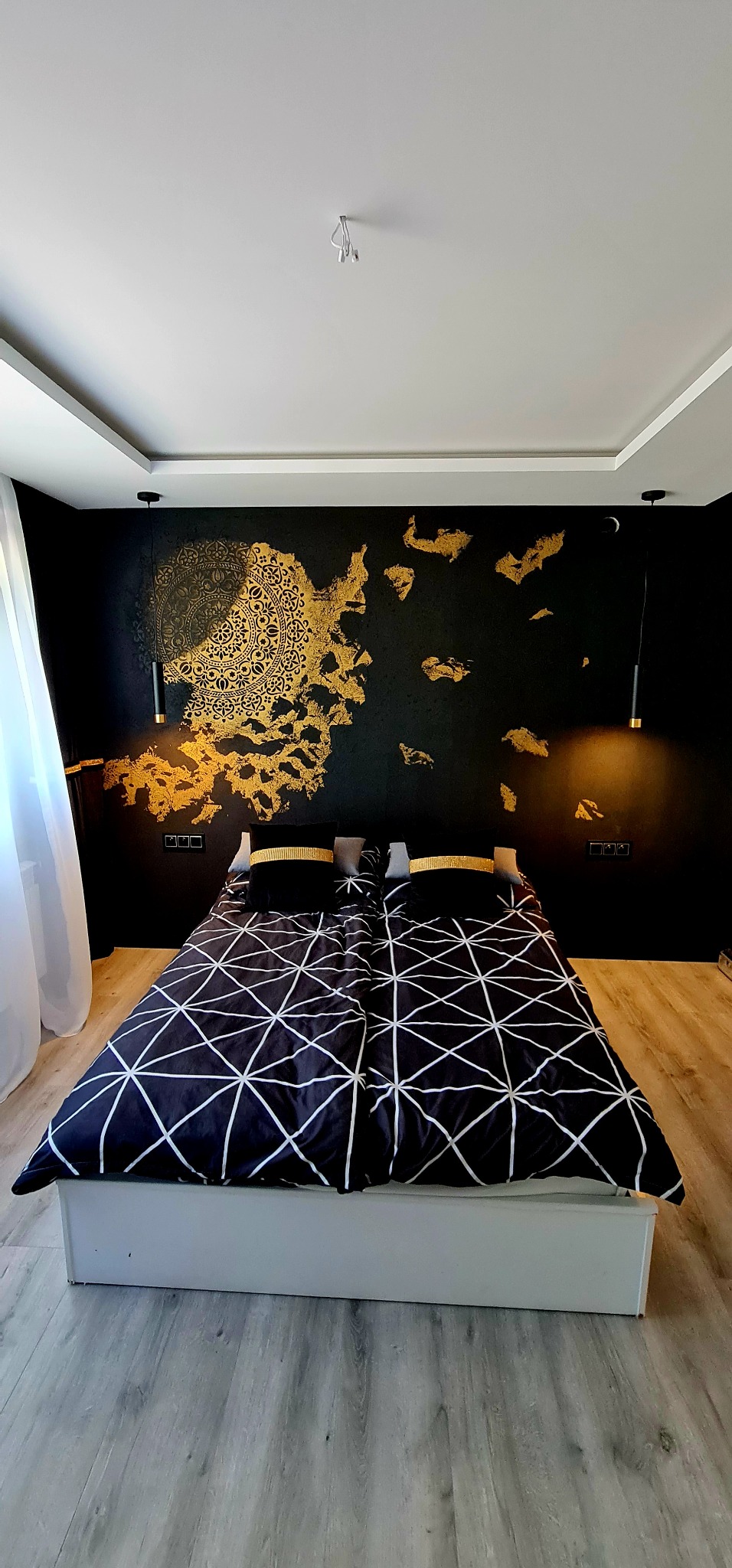 Sypialnia z czarną ścianą ozdobioną złotym, abstrakcyjnym wzorem, łóżko z geometryczną pościelą i czarnymi poduszkami z złotymi akcentami, wiszące lampy po bokach łóżka.