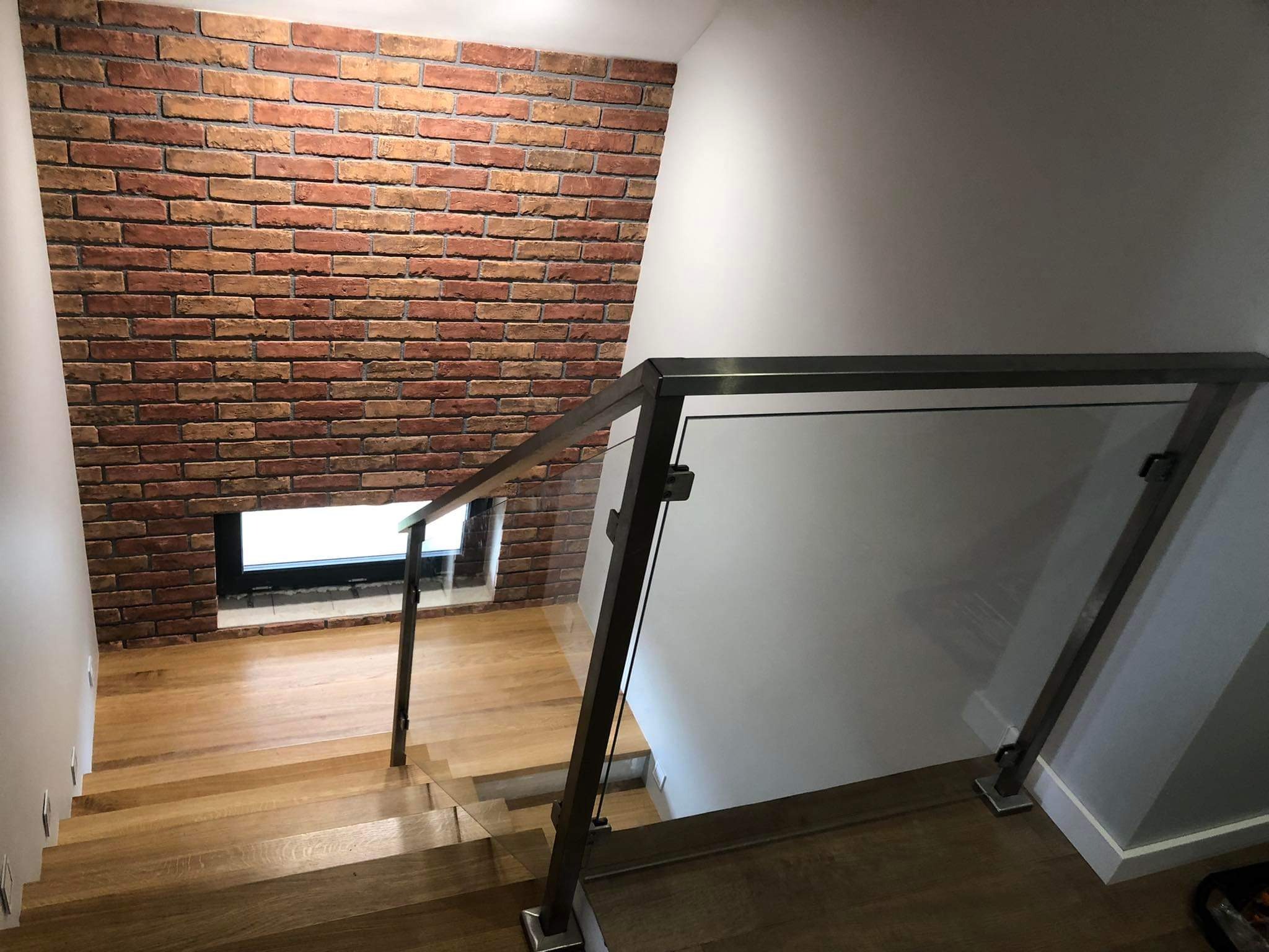 Schody drewniane z balustradą ze szkła i metalu, ściana z cegły dekoracyjnej w tle, widok z góry.