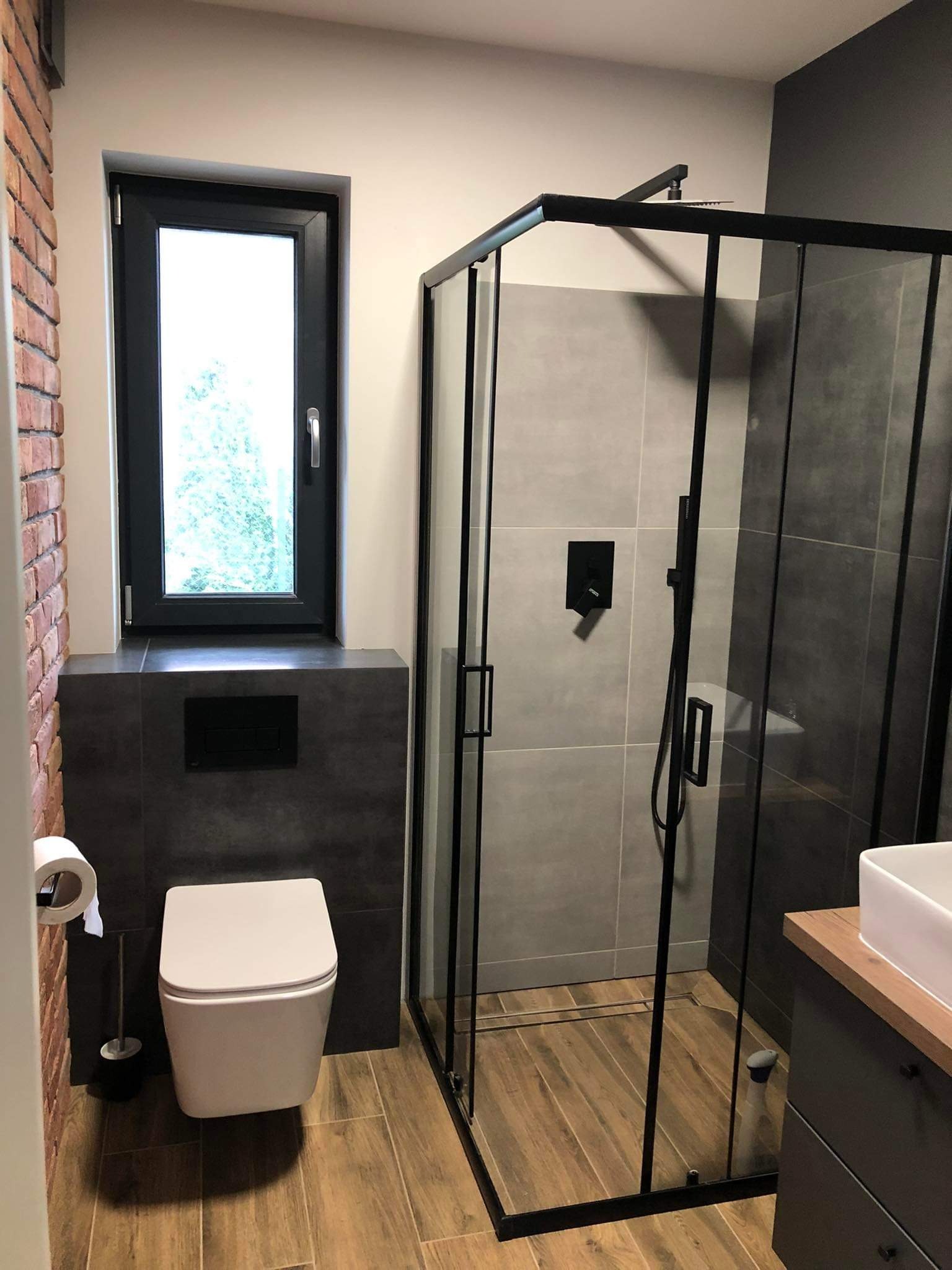 Nowoczesna łazienka z kabiną prysznicową typu walk-in z czarnymi profilami, podwieszaną toaletą i ścianą z cegły dekoracyjnej, drewnopodobna podłoga.
