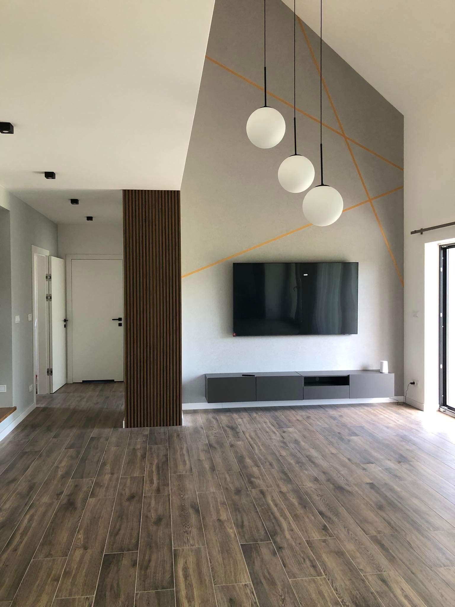 Nowoczesny salon z drewnopodobną podłogą, szarą ścianą z geometrycznym wzorem w kolorze złota, wiszącymi lampami kulami i panelem dekoracyjnym z pionowych listew.