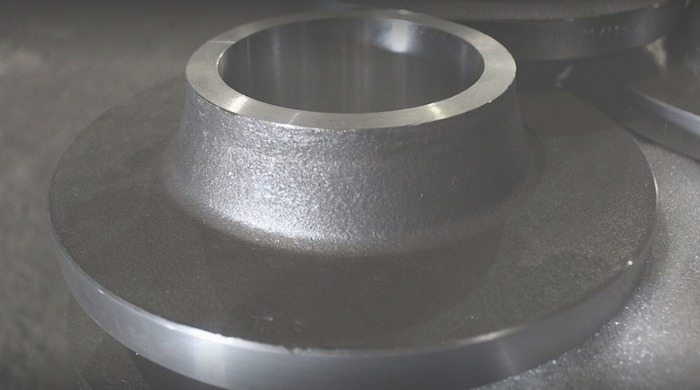 Szara, metalowa kształtka kołnierzowa z otworem pośrodku, leżąca na szarej powierzchni. Widoczna faktura obróbki.