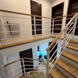 Wnętrze domu z drewnianymi schodami i białą balustradą z drewnianym poręczą, widok na antresolę z grzejnikiem i trzema parami ciemnych drzwi z poziomymi wstawkami.