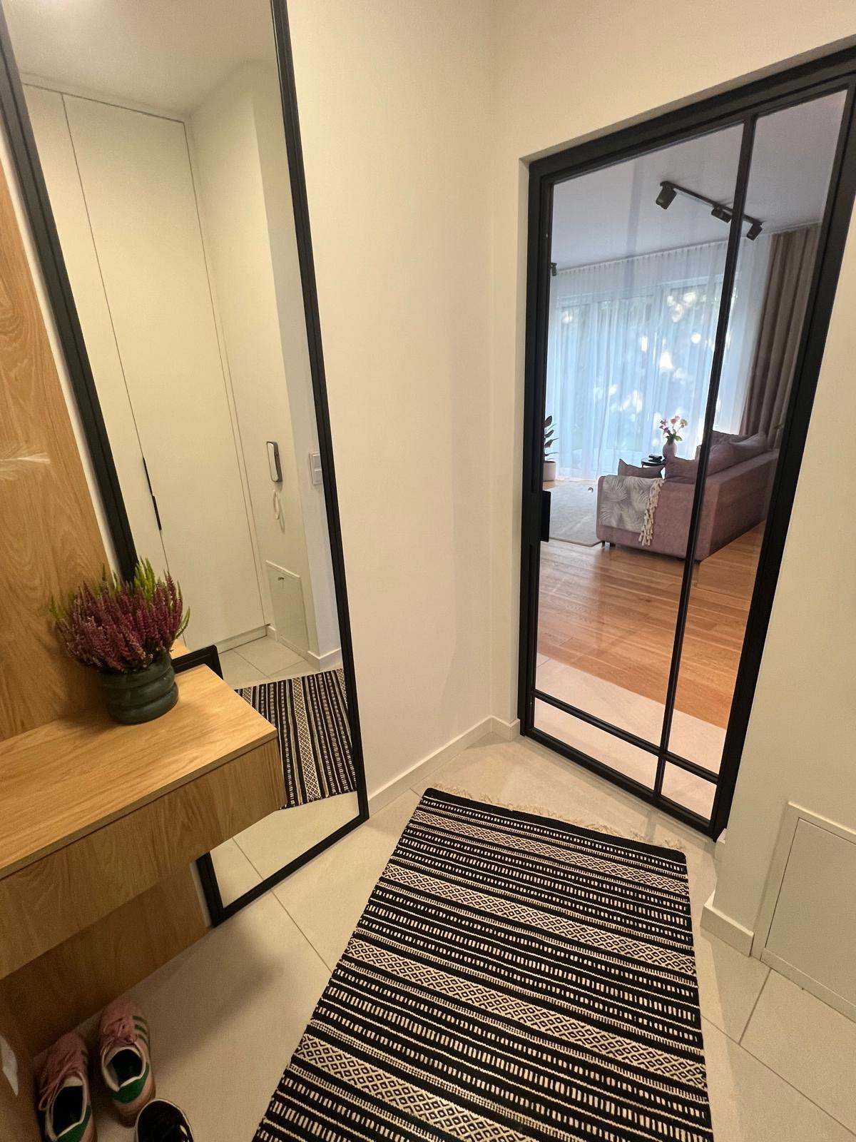 Nowoczesny przedpokój z lustrem w czarnej ramie i drzwiami szklanymi z czarnymi szprosami, dywanik w czarno-białe pasy na jasnych płytkach podłogowych.