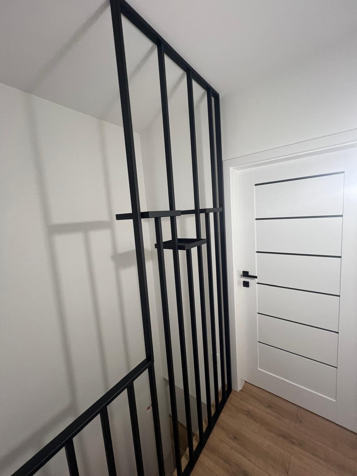 Nowoczesna czarna barierka schodowa z pionowych prętów, z półkami, obok białych drzwi z poziomymi wgłębieniami, na drewnianej podłodze.
