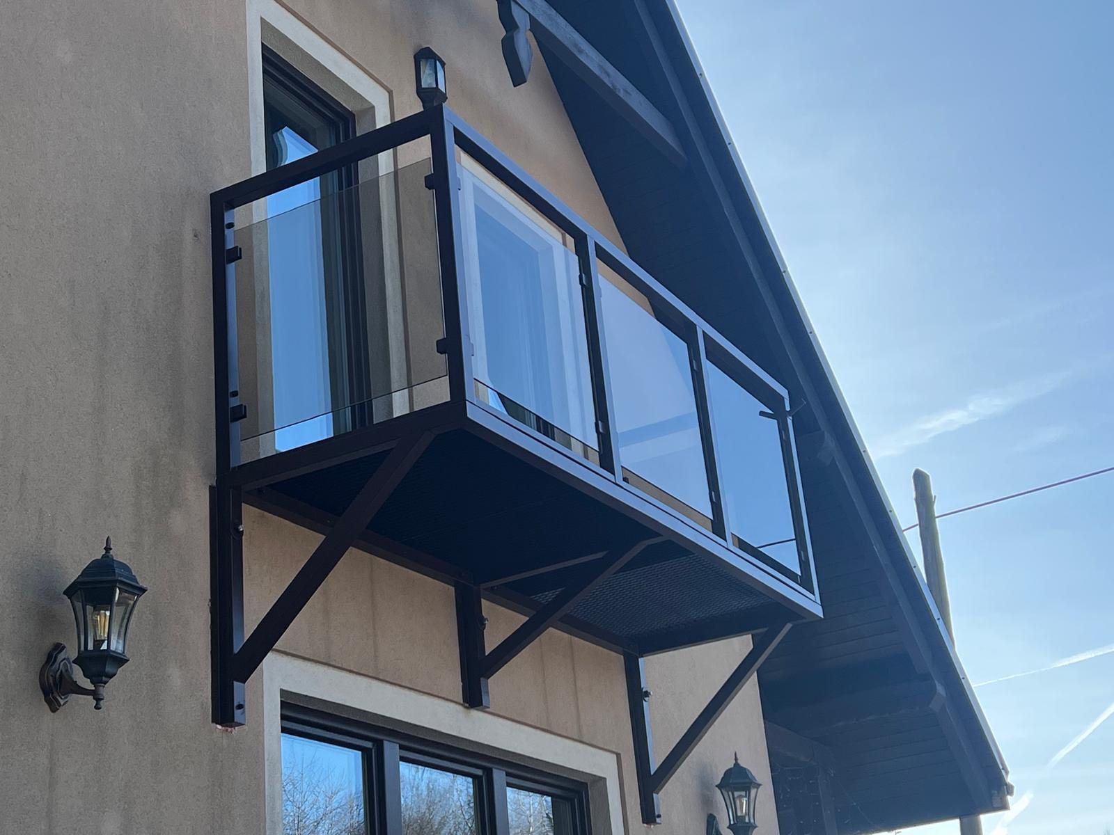 Zewnętrzny balkon z czarną, metalową konstrukcją i szklanymi panelami, zamontowany na fasadzie budynku z oknami i lampami ściennymi.