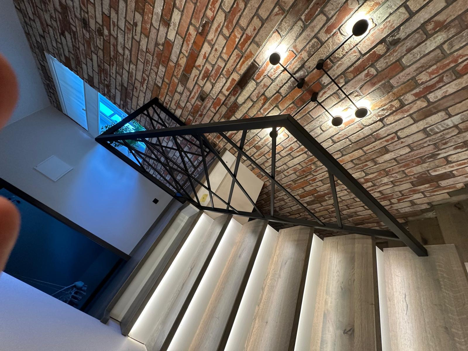 Nowoczesna balustrada z czarnej stali z geometrycznym wzorem w domu w Modlnicy, widok od dołu na schody z podświetleniem LED i ceglany sufit z designerską lampą.