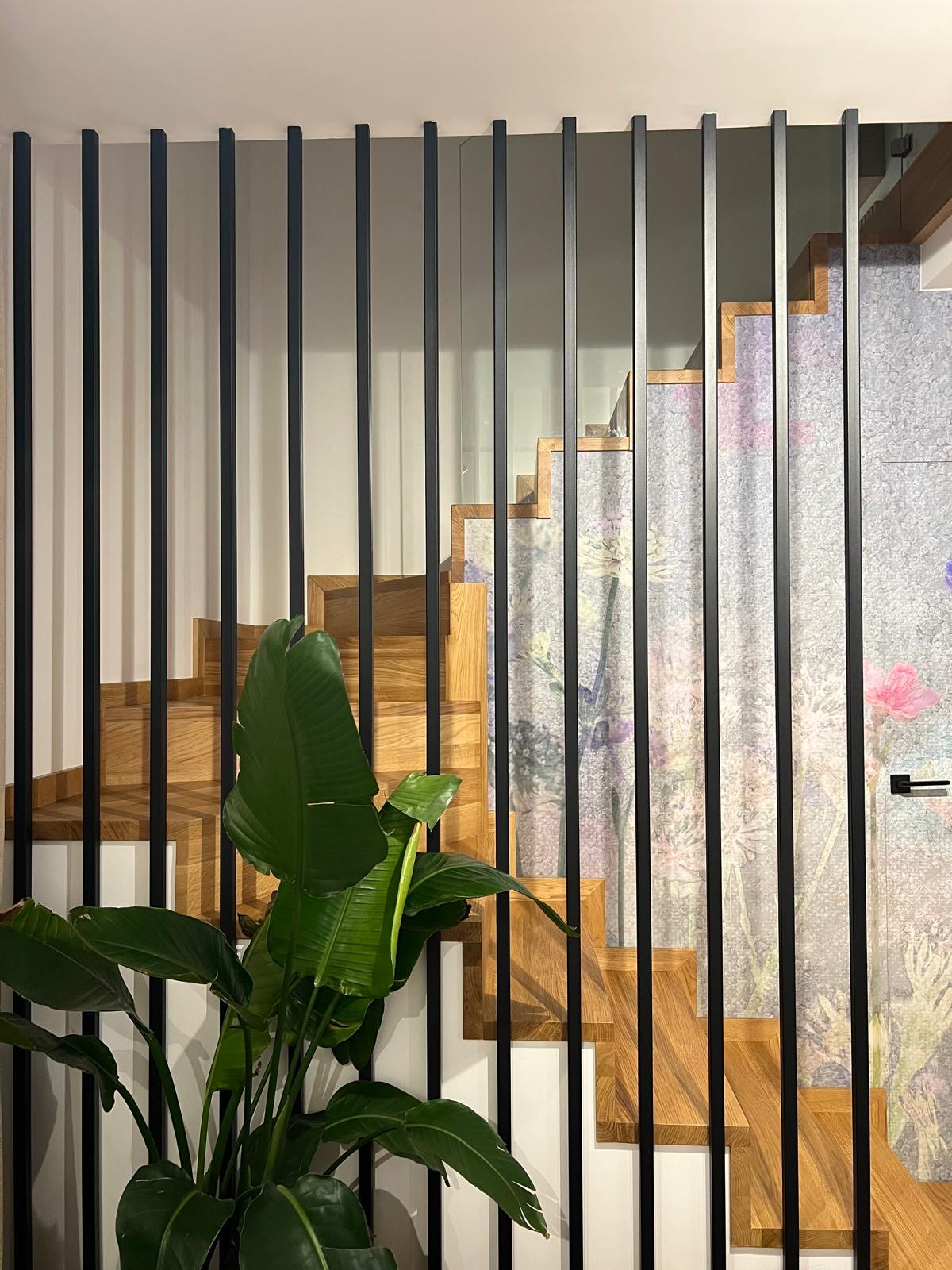 Nowoczesne schody drewniane z czarnymi, pionowymi prętami balustrady, częściowo przesłonięte rośliną doniczkową, z tapetą w kwiaty w tle.