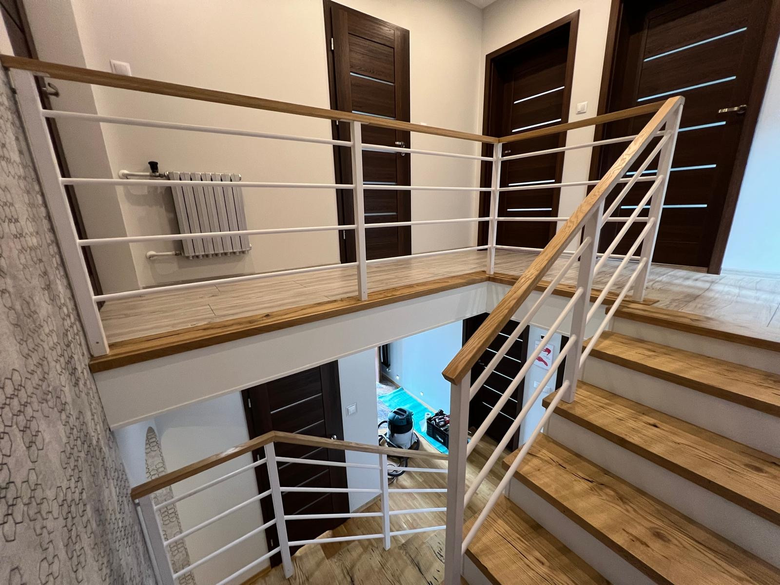 Wnętrze domu z drewnianymi schodami i białą balustradą z drewnianym poręczą, widok na antresolę z grzejnikiem i trzema parami ciemnych drzwi z poziomymi wstawkami.