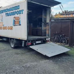 Ciężarówka z otwartą rampą załadowana meblami, zaparkowana na żwirowym podwórku, z rowerami opartymi o garaż.