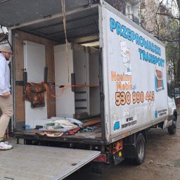 NosimyMeble - Mężczyzna w czapce i jasnych spodniach wchodzi do otwartego samochodu dostawczego z napisem 'Przeprowadzki Transport' załadowanego szafkami i zabezpieczonego pasami, w tle budynek mieszkalny i drzewa.