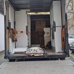 NosimyMeble - Otwarta paka samochodu dostawczego wypełniona meblami i zabezpieczonymi przedmiotami, widoczny mężczyzna w beżowej czapce i białej bluzie, prawdopodobnie kierowca, stojący na rampie załadunkowej.