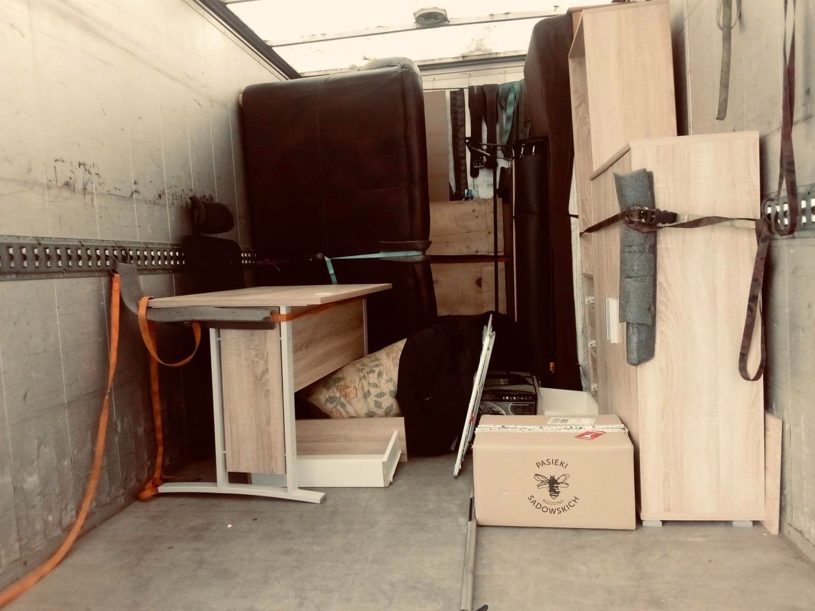 Wnętrze furgonetki załadowane meblami: biurko z jasnego drewna, ciemna sofa, szafki zabezpieczone pasami, karton z logo pszczoły.