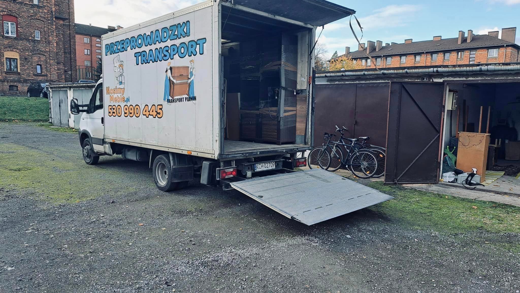 Ciężarówka z otwartą rampą załadowana meblami, zaparkowana na żwirowym podwórku, z rowerami opartymi o garaż.