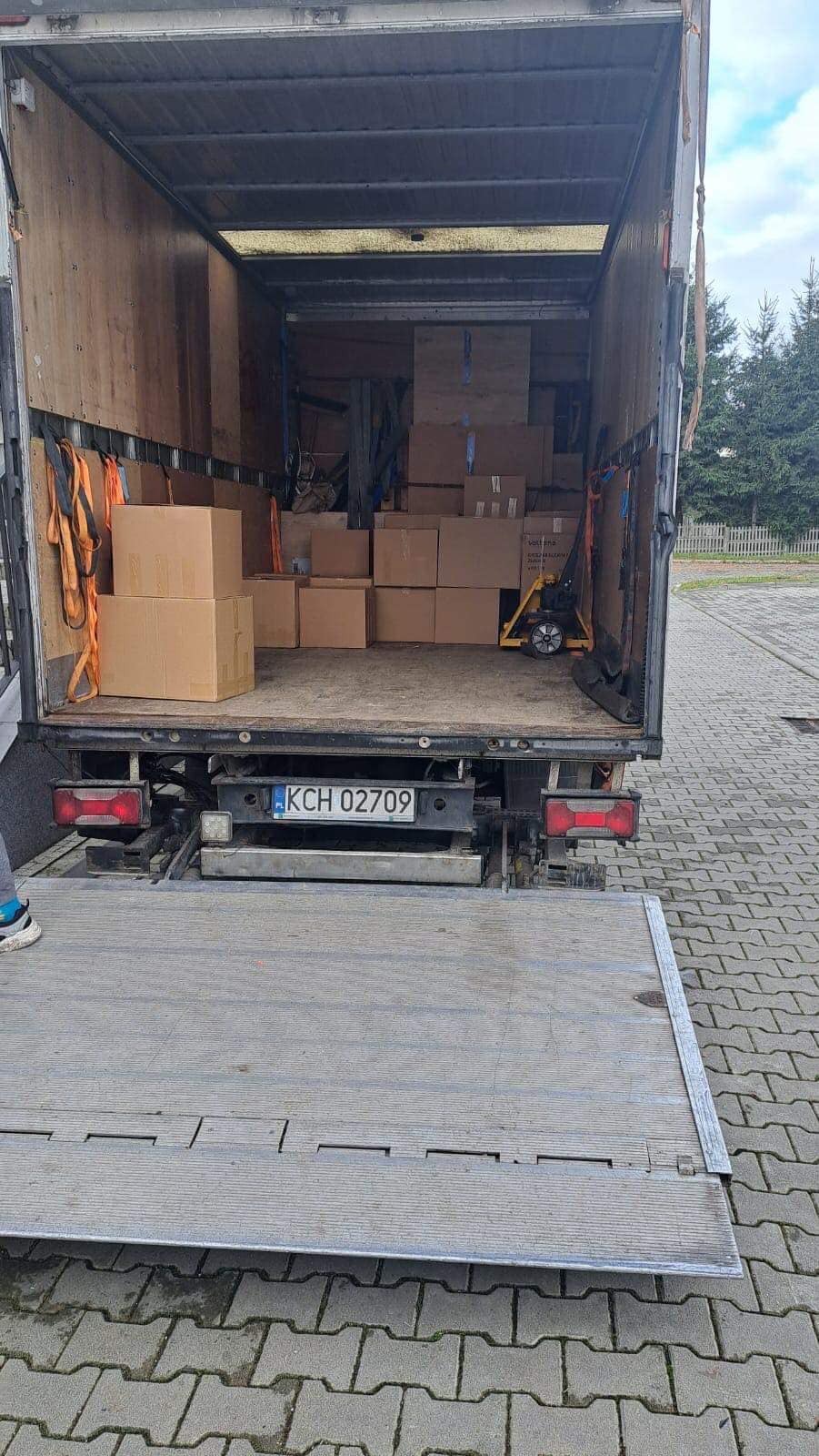 Otwarta przestrzeń ładunkowa ciężarówki wypełniona kartonami, zabezpieczona pasami transportowymi, z widoczną tablicą rejestracyjną KCH 02709 i podnośnikiem hydraulicznym