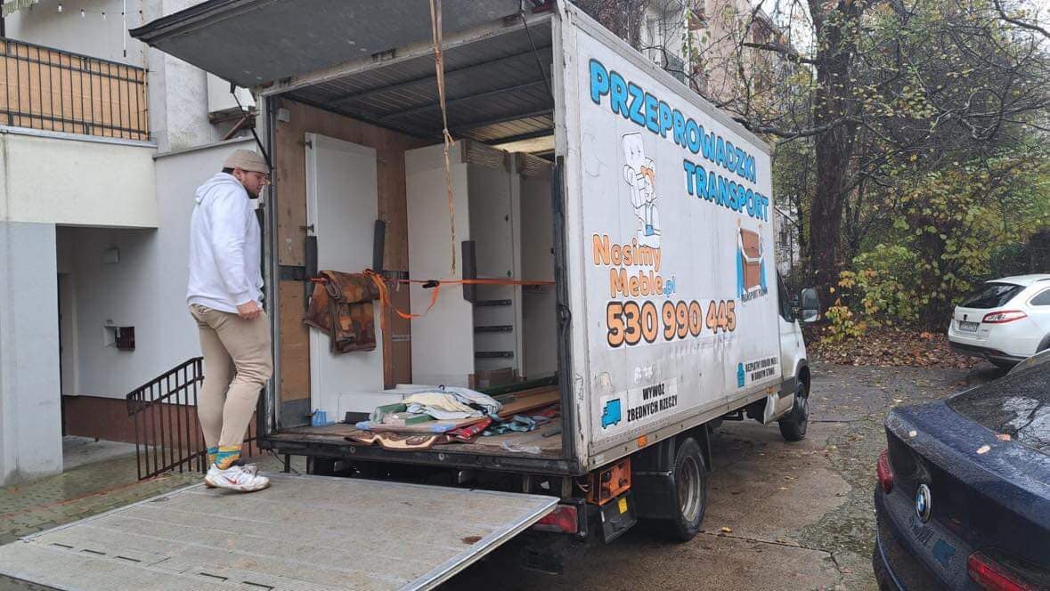 Mężczyzna w czapce i jasnych spodniach wchodzi do otwartego samochodu dostawczego z napisem 'Przeprowadzki Transport' załadowanego szafkami i zabezpieczonego pasami, w tle budynek mieszkalny i drzewa.