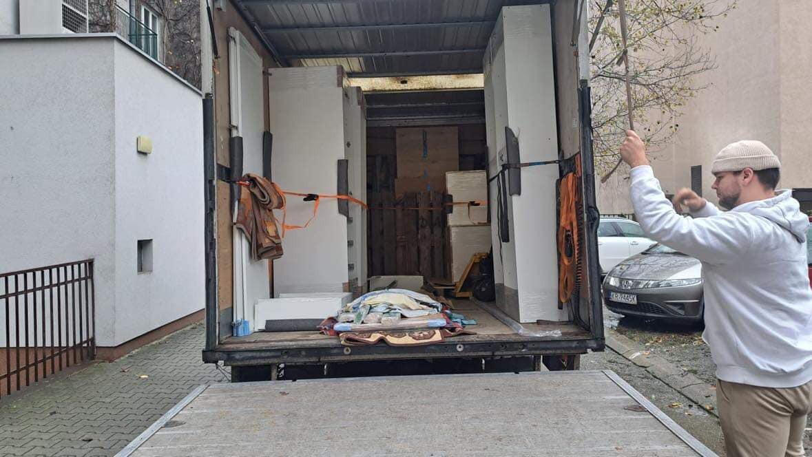 Otwarta paka samochodu dostawczego wypełniona meblami i zabezpieczonymi przedmiotami, widoczny mężczyzna w beżowej czapce i białej bluzie, prawdopodobnie kierowca, stojący na rampie załadunkowej.