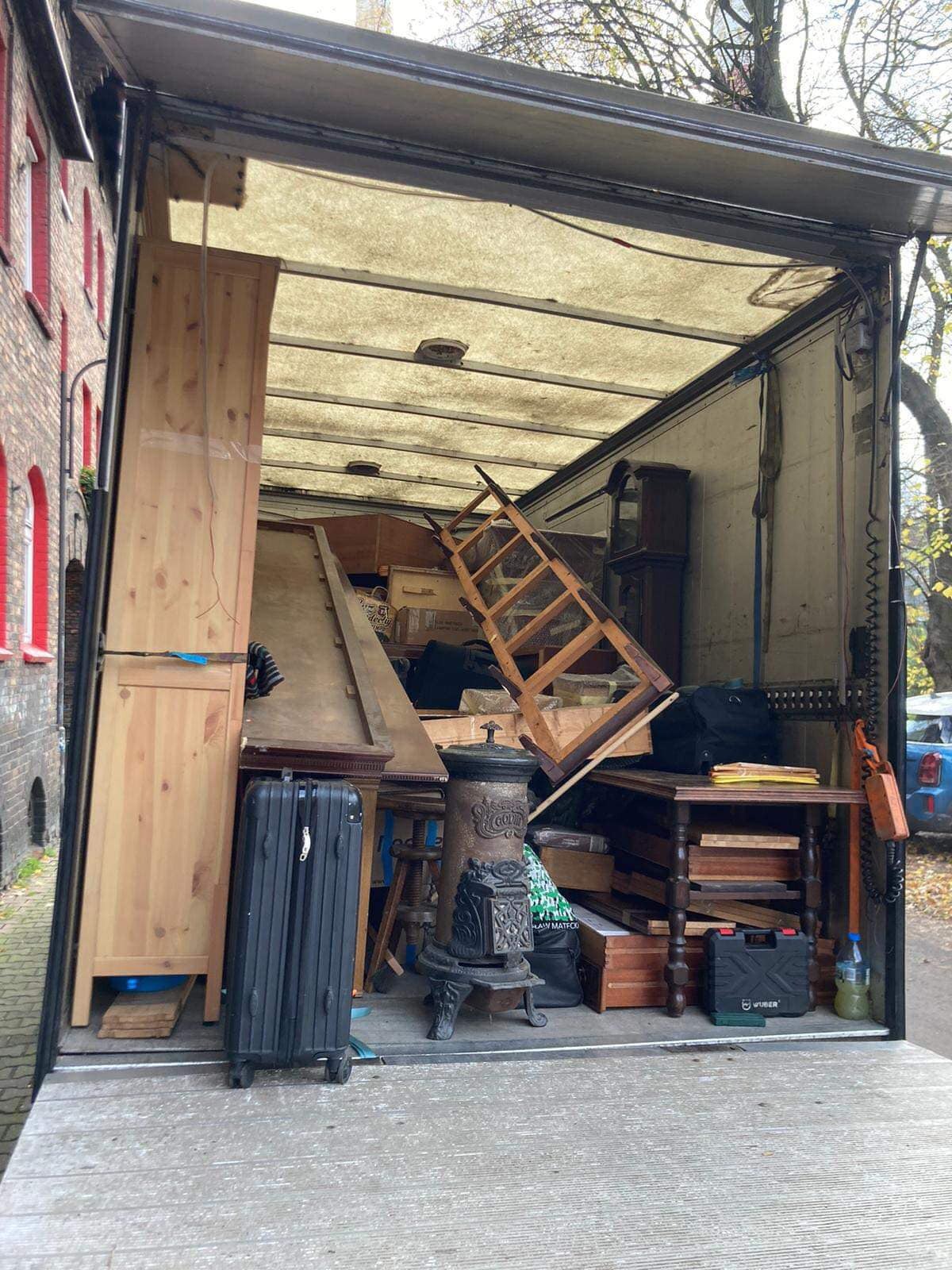 Wnętrze busa transportowego załadowane meblami i osobistymi rzeczami podczas przeprowadzki, widoczny piec żeliwny, szafa z jasnego drewna i czarna walizka na kółkach.