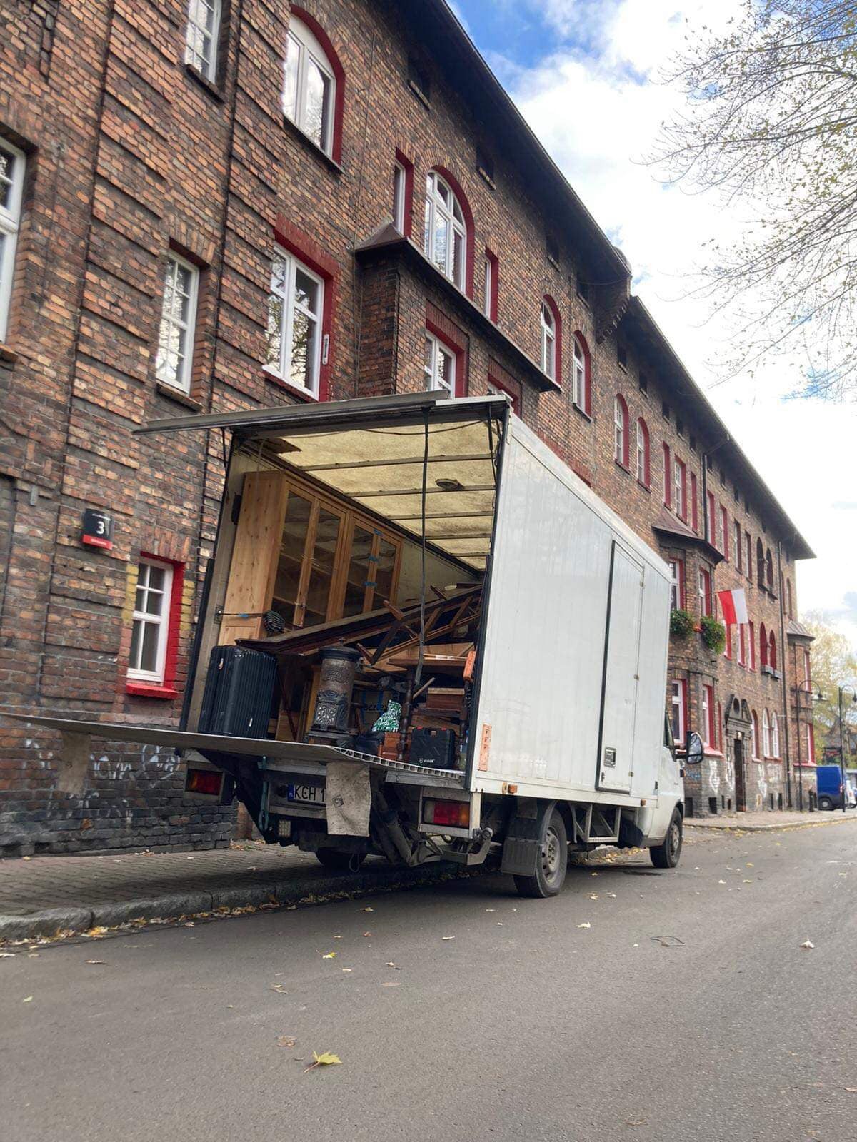 Otwarty furgon załadowany meblami i bagażami, zaparkowany na ulicy przed ceglanym budynkiem z czerwonymi obramowaniami okien.