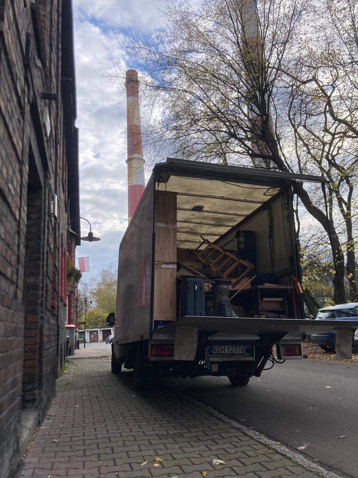 Otwarty tył brudnego samochodu dostawczego załadowanego meblami i bagażami na tle ceglanego budynku i komina, widok z chodnika.