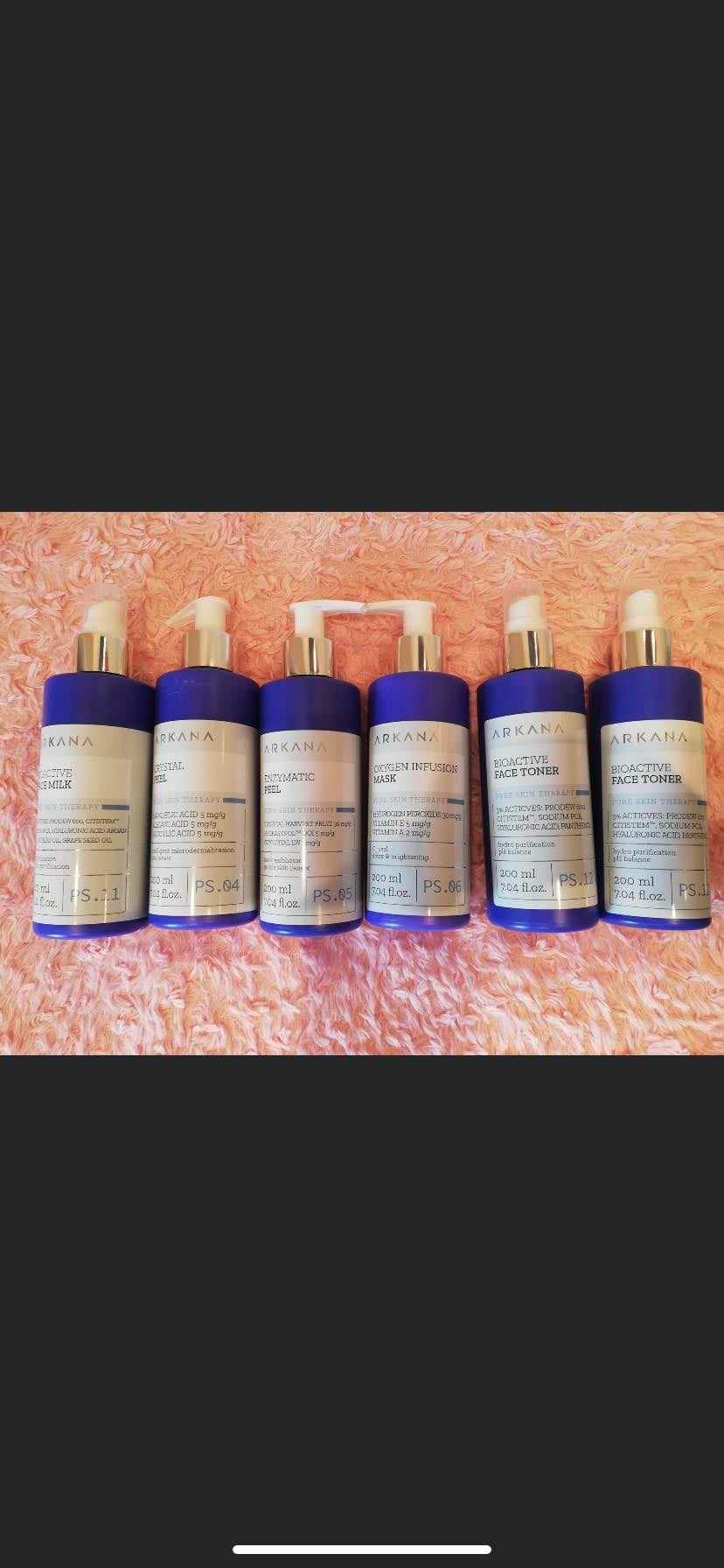 Rząd sześciu butelek kosmetyków marki Arkana z pompkami i ciemnofioletowymi nakrętkami, ułożonych na puszystym, różowym tle. Widoczne różne rodzaje produktów: mleczko, peeling, maska i tonik...