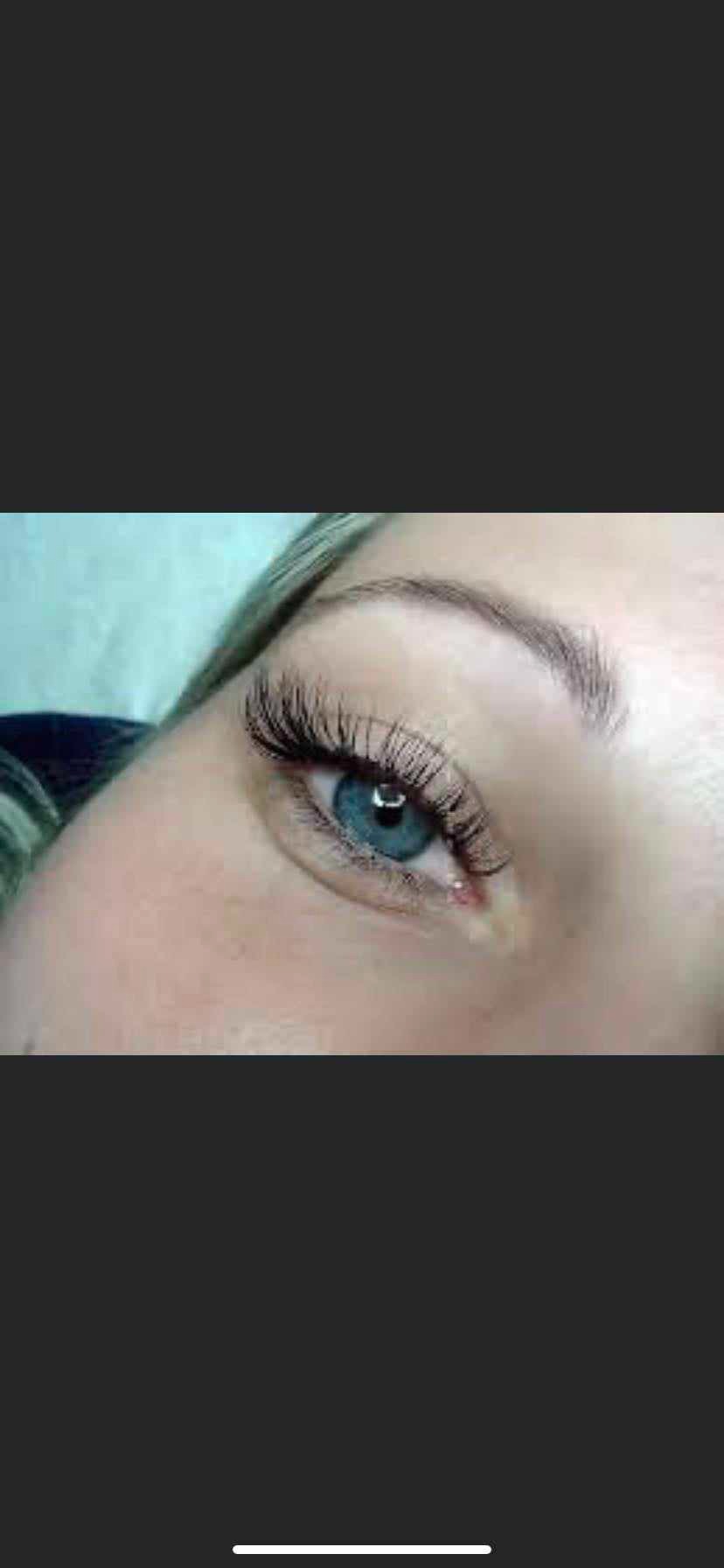 Zbliżenie na kobiece oko z niebieską tęczówką i perfekcyjnie przedłużonymi, czarnymi rzęsami; widoczny delikatny makijaż brwi.