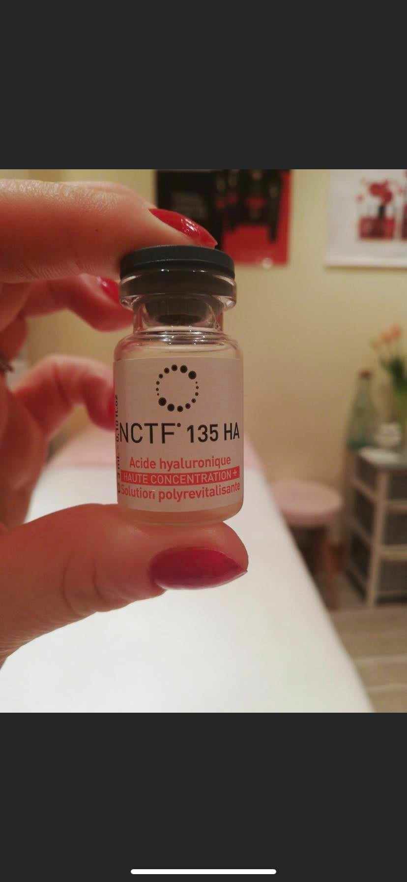 Fiolka z preparatem NCTF 135 HA, trzymana w palcach dłoni z czerwonym manicure, na rozmytym tle gabinetu kosmetycznego.