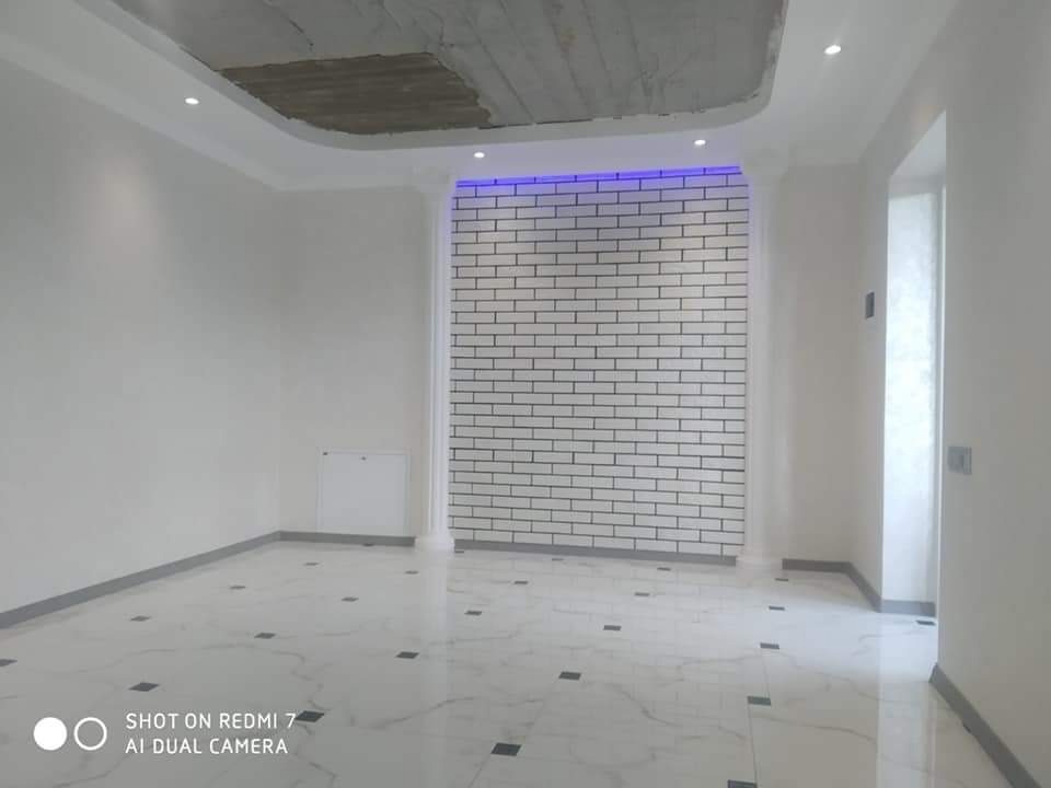 Wnętrze pokoju z białymi ścianami, marmurową podłogą z czarnymi akcentami i fragmentem sufitu z widoczną instalacją, oświetlone punktowymi lampami i niebieskim światłem nad ścianą z białej cegły.